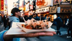 A close-up of a hand holding a wooden tray of takoyaki, Japanese octopus balls topped with bonito flakes, mayonnaise, and green onions. Toothpicks are inserted for easy eating. In the background, a lively street scene with people walking and storefronts with Japanese signage suggests a bustling food market atmosphere.