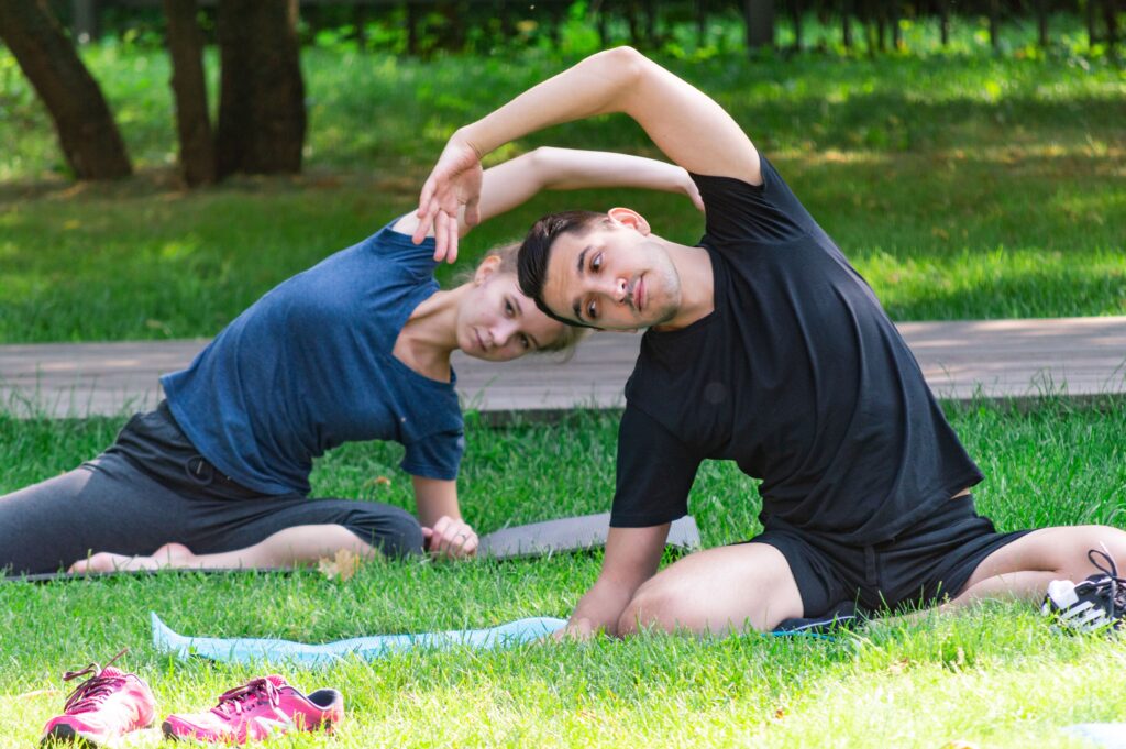 A man and woman sitting on grass outdoors, stretching sideways on yoga mats, creating a peaceful and reflective scene — symbolising the journey of a time traveler moving through seasons.