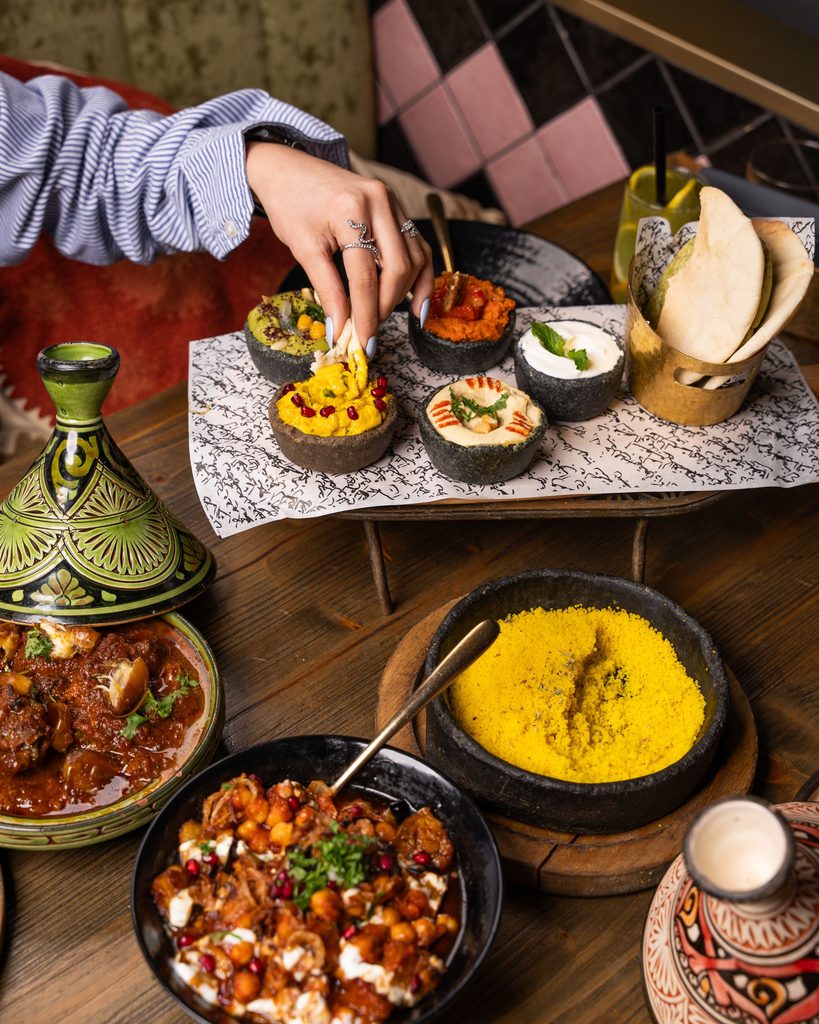 A woman’s hand reaching for food on a traditional Moroccan table, showing warmth, hospitality, and shared meals within the desert palette.