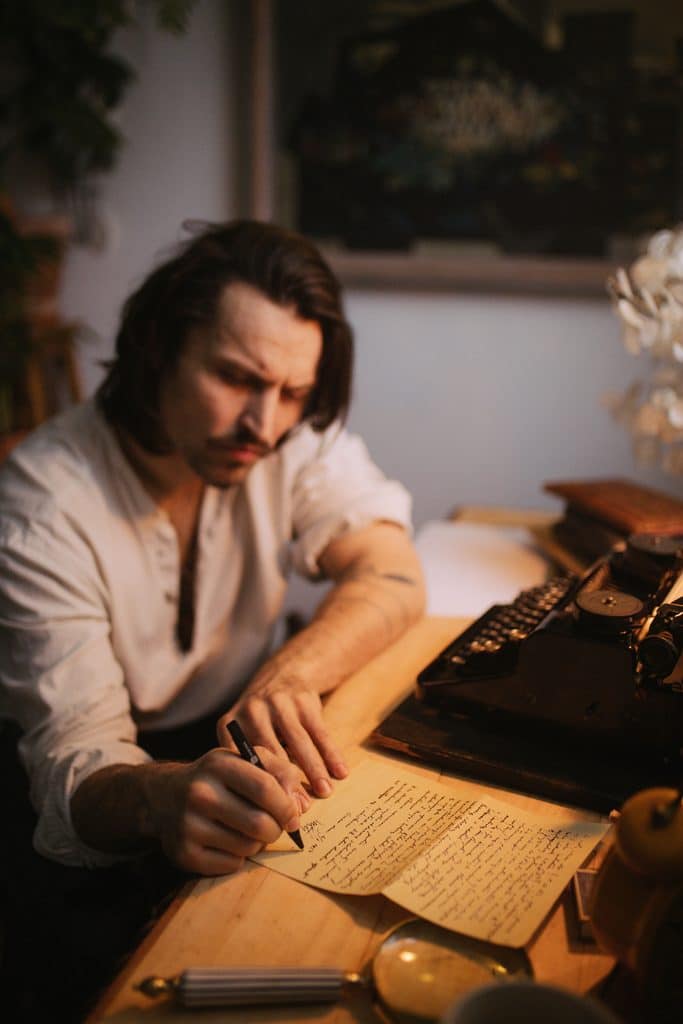 A man sitting at a table writing on paper in soft natural light, creating a quiet, reflective Nordic‑style interior scene.